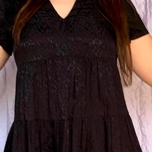 May include: A black short-sleeved dress with a V-neck and a tiered skirt. The dress features a textured pattern across the fabric. The dress is a solid black colour.