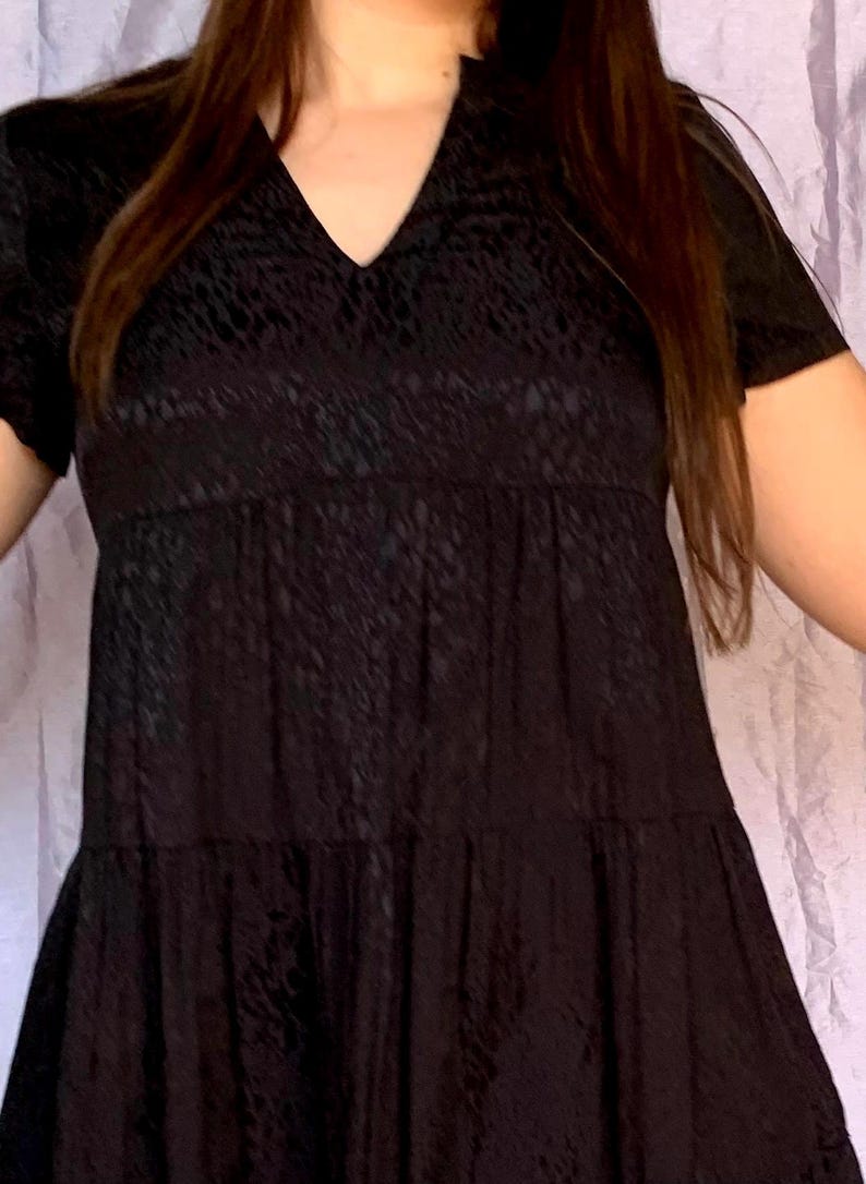 May include: A black short-sleeved dress with a V-neck and a tiered skirt. The dress features a textured pattern across the fabric. The dress is a solid black colour.