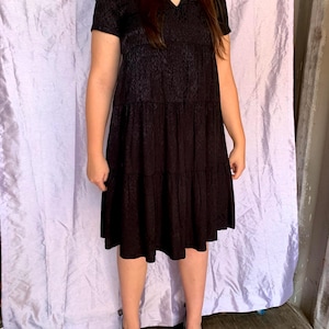 May include: A black, short-sleeved dress with a V-neck and tiered skirt. The dress features a subtle, textured pattern and is worn with black heels. The dress is knee-length and appears to be made of a lightweight fabric.
