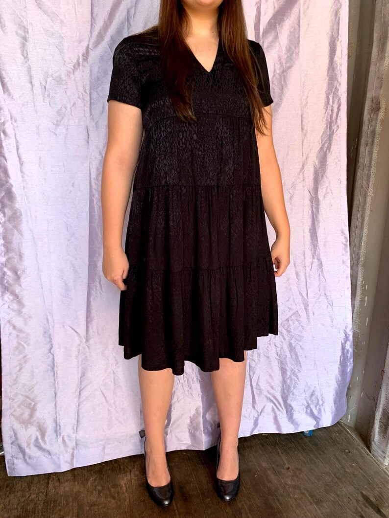 May include: A black, short-sleeved dress with a V-neck and tiered skirt. The dress features a subtle, textured pattern and is worn with black heels. The dress is knee-length and appears to be made of a lightweight fabric.