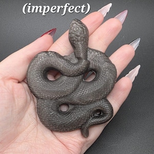 May include: A dark grey silver sheen obsidian snake sculpture, weighing 60g. The coiled serpent has detailed scales and a raised head. The image includes text: "silver sheen obsidian 60g (imperfect)".