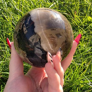 May include: A polished, spherical crystal ball with a reflective surface, displaying a mix of dark brown and clear tones. The sphere is held against a backdrop of green grass, creating a contrast of textures and colors. The crystal ball is a decorative item.