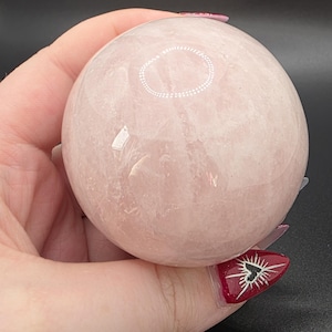 May include: A polished, spherical rose quartz crystal with a smooth, translucent pale pink surface. The stone features natural internal inclusions and subtle variations in opacity, reflecting light across its rounded, symmetrical form.