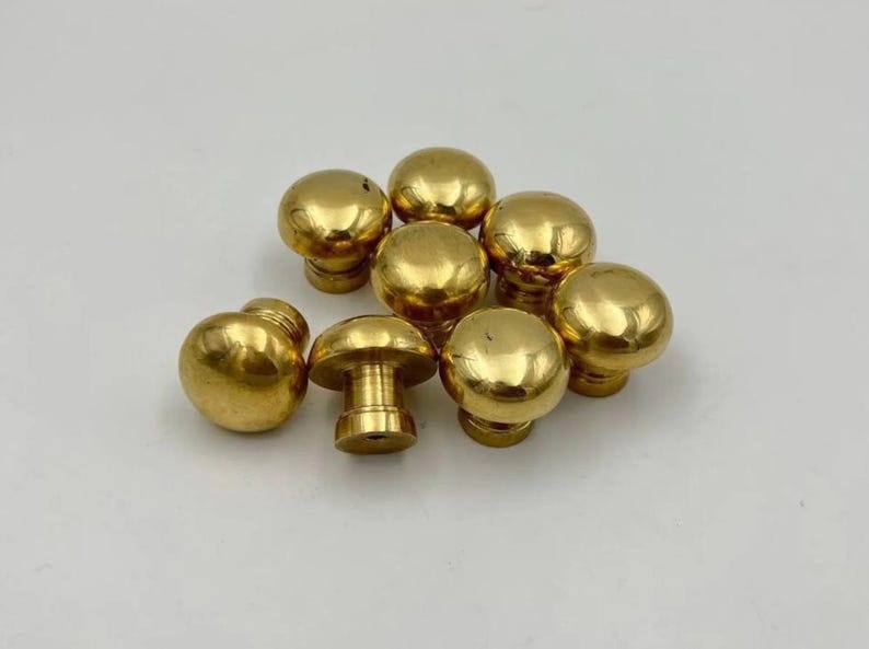 May include: A collection of eight small, polished gold-toned metal knobs. Each knob features a smooth, rounded, mushroom-shaped head attached to a cylindrical base. The metallic surface is reflective, showing highlights and shadows across the set.