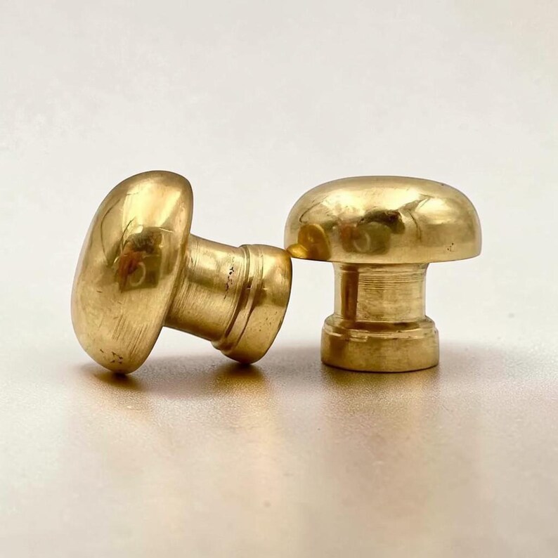 May include: Two polished brass cabinet knobs with a smooth, rounded mushroom-shaped head and a cylindrical base. The metal surface has a reflective, golden-yellow finish with subtle variations in tone, showing a classic hardware design.