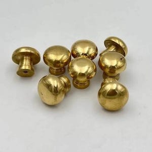 May include: A collection of seven polished brass cabinet knobs, each featuring a smooth, rounded mushroom-shaped head and a cylindrical base with a central screw hole, displaying a reflective metallic gold finish across all pieces.