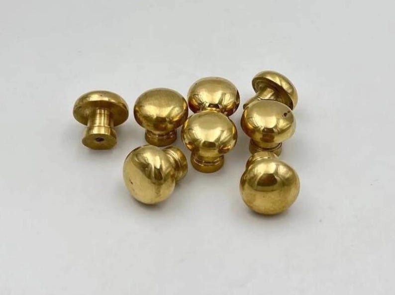 May include: A collection of seven polished brass cabinet knobs, each featuring a smooth, rounded mushroom-shaped head and a cylindrical base with a central screw hole, displaying a reflective metallic gold finish across all pieces.