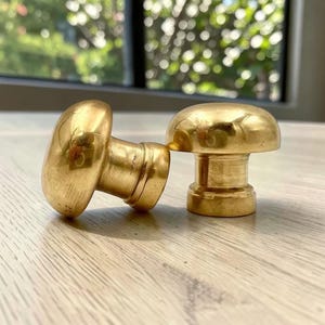 May include: Two polished brass cabinet knobs with a smooth, rounded mushroom-shaped head and a cylindrical base. The metal surface has a reflective, golden finish that catches the light, showing a simple, functional design for furniture hardware.