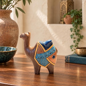 May include: A brown camel figurine with a textured surface, featuring a decorative saddle blanket in shades of blue, orange, and yellow. The saddle blanket is adorned with geometric patterns, including triangles, lines, and a small star shape.