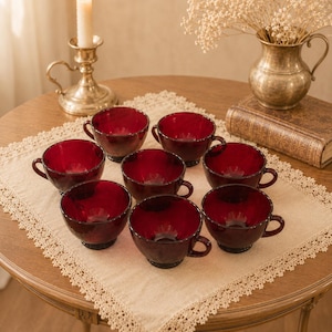 May include: A set of eight deep red glass teacups with rounded bowls, short stems, and small side handles. The cups are arranged in a cluster on a square beige lace-trimmed fabric mat, showcasing a translucent, dark ruby-colored glass finish.