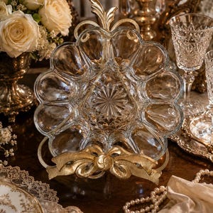 May include: A clear glass serving dish with a gold-colored metal bow-shaped accent. The dish has a starburst design in the center and multiple rounded compartments. The gold accent is at the base of the dish. The dish is surrounded by other decorative items.