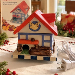 May include: An electric simmering potpourri pot shaped like a house with a red roof, white walls, and blue trim. The front features a yellow bird in a circular cutout, a small orange porch, and a stack of brown logs. It includes a white power cord.