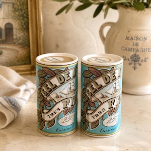 May include: Two cylindrical containers featuring a light blue background with a graphic illustration of a sailing ship, green leaves, and the text "SEL DE MER TRES FIN 250g Cuisine". The containers have a white top rim and a tan circular lid.