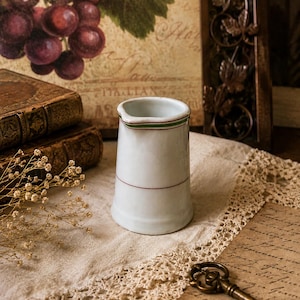 May include: A white ceramic creamer pitcher featuring a simple design with a thin green and red horizontal stripe around the rim and a single thin red horizontal stripe encircling the lower body, with a small pouring spout at the top edge.