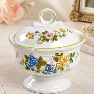 May include: A white porcelain sugar bowl with a matching lid featuring a ring-shaped handle. The bowl and lid are decorated with colorful floral patterns in blue, yellow, and pink, accented by green leaves and a thin green rim along the edges.