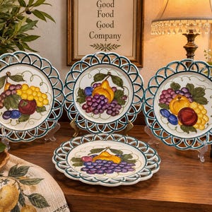 May include: A set of four round ceramic plates featuring a decorative lattice-style openwork border in dark teal. Each plate displays a hand-painted central motif of assorted fruits, including yellow and purple grapes, pears, and plums on a white base.