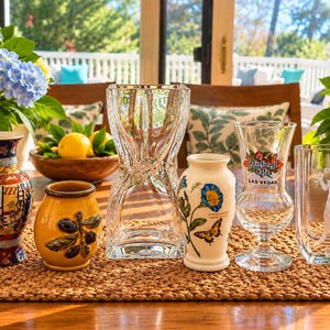 May include: A collection of six decorative items: a colorful patterned vase, a yellow ceramic pot with olive branch relief, a clear hourglass-shaped glass vase, a floral vase with a butterfly, a Rainforest Cafe Las Vegas glass, and a clear glass vase with an etched figure.
