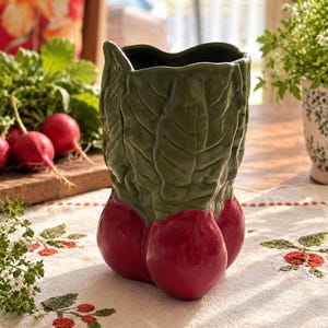 May include: A ceramic vase shaped like a bunch of radishes, featuring a glossy red base representing the root vegetables and a textured green top sculpted to resemble the leafy greens of the plant, with an open top for holding items.