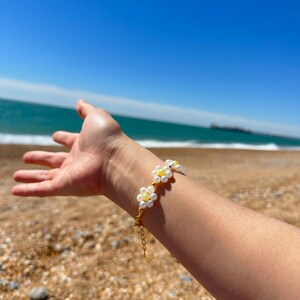 May include: A gold-toned bracelet featuring daisy-shaped beads with white petals and yellow centres. The bracelet is worn on a wrist, set against a backdrop of a beach and blue sea.
