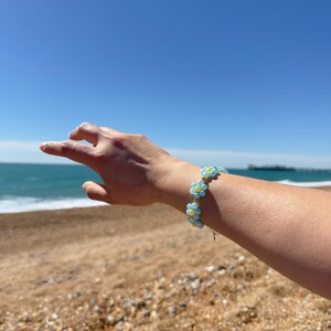 May include: A bracelet made of light blue and yellow daisy flower beads. The bracelet is worn on a wrist against a beach and blue sky. The jewelry is a delicate piece. The bracelet is a pretty accessory.
