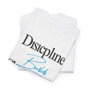 May include: Two white t-shirts are folded, with the top one printed with the words "Discipline Builds" in black and blue. The shirts are made of a soft material, likely cotton, and are ready to wear.