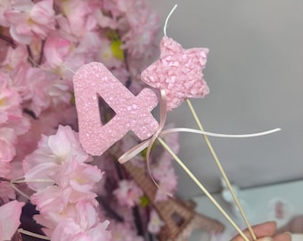 Puede incluir: Una vela con el número cuatro en rosa claro y una vela con forma de estrella en rosa claro, ambas con una superficie texturizada y cristalina. Cada vela está montada en un palito de madera, unidos por una cinta rosa claro.