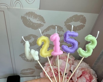 Puede incluir: Cinco velas numéricas individuales en colores pastel que incluyen blanco, amarillo, rosa, morado y verde claro. Cada vela tiene forma de dígito y está montada sobre un fino palito de madera, con una pequeña mecha blanca en la parte superior.
