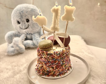 Puede incluir: Un pastel pequeño cubierto de chispas multicolores, coronado con tres velas encendidas con forma de elefante, el número uno y un osito de peluche, atado con una cinta blanca. Un pulpo de peluche azul claro se encuentra junto al pastel en un plato.