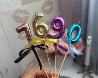 Puede incluir: Cuatro velas numéricas metálicas sobre palitos de madera, que representan el 1, 6, 9 y 0. El 1 es oro rosa con cinta marrón, el 6 es dorado con cinta amarilla, el 9 es morado con cinta morada y el 0 es azul con cinta azul claro.