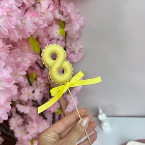 May include: A pale yellow number eight candle topper shaped like a balloon, mounted on a thin wooden stick. The candle is decorated with a bright yellow grosgrain ribbon tied in a bow just below the number, creating a festive cake decoration.