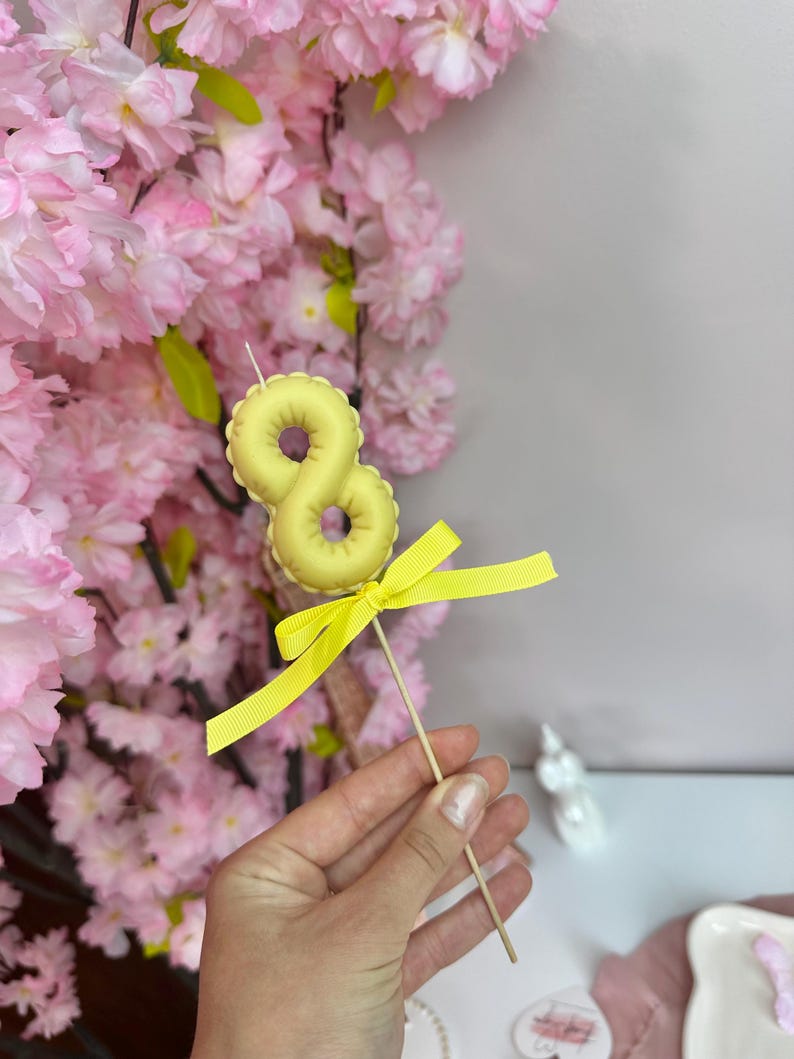 May include: A pale yellow number eight candle topper shaped like a balloon, mounted on a thin wooden stick. The candle is decorated with a bright yellow grosgrain ribbon tied in a bow just below the number, creating a festive cake decoration.