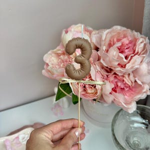 May include: A brown number three candle topper with a textured, balloon-like appearance. The candle is attached to a thin wooden stick and features a small, shimmering gold ribbon tied just below the number, with a single white wick on top.