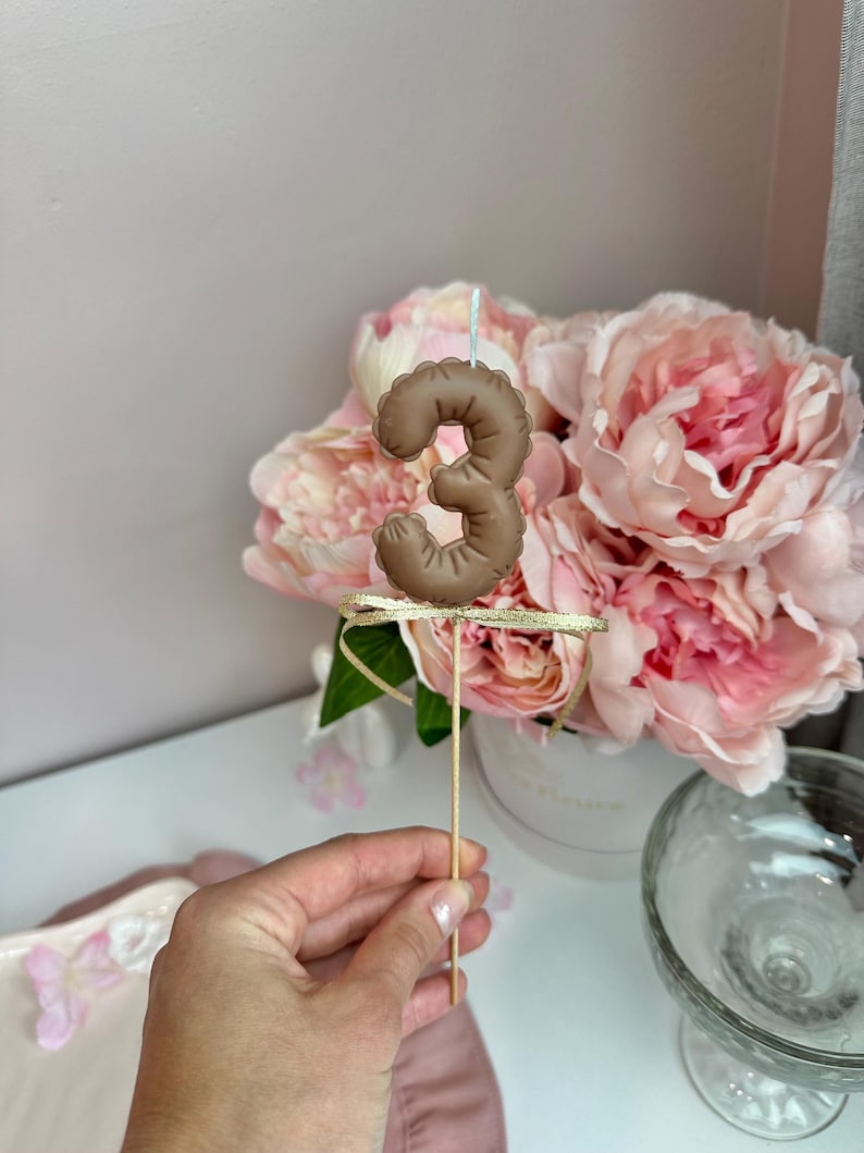 May include: A brown number three candle topper with a textured, balloon-like appearance. The candle is attached to a thin wooden stick and features a small, shimmering gold ribbon tied just below the number, with a single white wick on top.