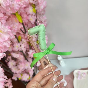May include: A light green number seven candle topper with a scalloped edge, tied with a bright green ribbon on a wooden stick. The candle features a small white wick at the top and is designed for placement on a cake or other food item.