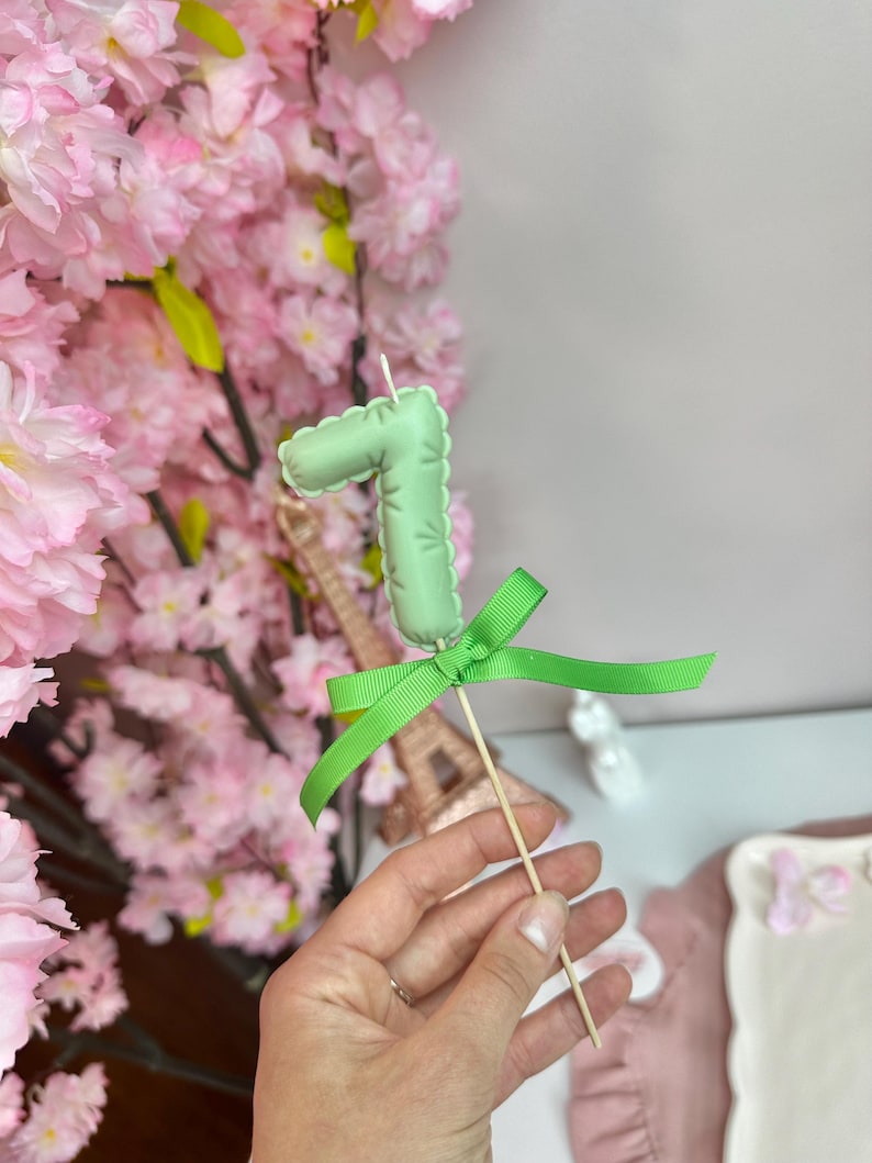 May include: A light green number seven candle topper with a scalloped edge, tied with a bright green ribbon on a wooden stick. The candle features a small white wick at the top and is designed for placement on a cake or other food item.