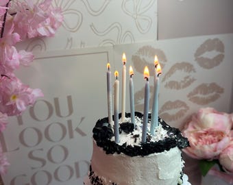 Puede incluir: Un pastel redondo pequeño con glaseado blanco, bordes negros y virutas de colores blanco y negro. Seis velas de cumpleaños finas y encendidas en tonos azules y blancos están insertadas sobre el pastel, que descansa en una base festoneada.