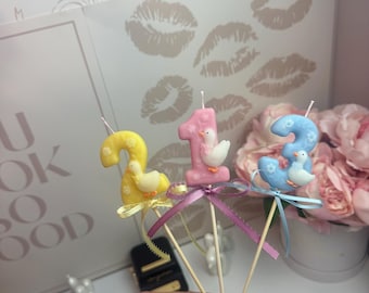 Puede incluir: Tres velas con forma de números sobre palitos de madera, que incluyen un dos amarillo, un uno rosa y un tres azul. Cada vela está decorada con una pequeña figura de pato blanco, detalles florales y una cinta de satén a juego atada debajo.