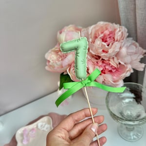 May include: A light green number seven cake topper made of fabric with a stitched edge, featuring a bright green ribbon tied in a bow at the base. The number is attached to a thin wooden stick for insertion into a cake or other food item.