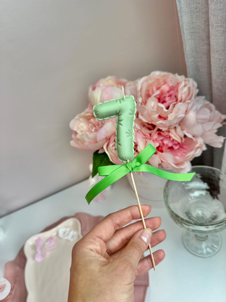 May include: A light green number seven cake topper made of fabric with a stitched edge, featuring a bright green ribbon tied in a bow at the base. The number is attached to a thin wooden stick for insertion into a cake or other food item.