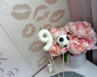 Puede incluir: Dos adornos para pasteles sobre finos palitos de madera. Uno es una vela blanca con forma de número nueve y superficie texturizada. El otro es un pequeño balón de fútbol redondo, de color blanco y negro, con un patrón hexagonal clásico.
