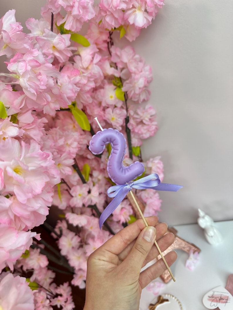 May include: A light purple number two candle topper shaped like a balloon, featuring a smooth finish and a small matching purple ribbon tied around the wooden stick base. The candle has a white wick at the top and a rounded, inflated appearance.