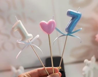 May include: Three cake toppers on wooden sticks: a white number one candle, a pink heart-shaped candle, and a blue number seven candle. Each candle features a small satin ribbon tied around the base of the stick, adding a decorative touch to the set.
