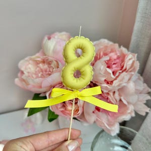May include: A pale yellow number eight candle shaped like a balloon, featuring a small white wick at the top and a bright yellow ribbon tied in a bow around the thin wooden stick base, positioned upright for display.