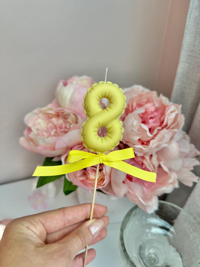 May include: A pale yellow number eight candle shaped like a balloon, featuring a small white wick at the top and a bright yellow ribbon tied in a bow around the thin wooden stick base, positioned upright for display.