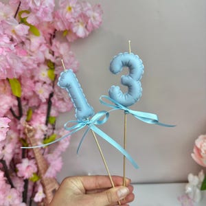 May include: Two light blue number candles, shaped like the digits one and three, are mounted on wooden sticks. Each candle is decorated with a matching light blue satin ribbon tied in a bow around the base of the number.