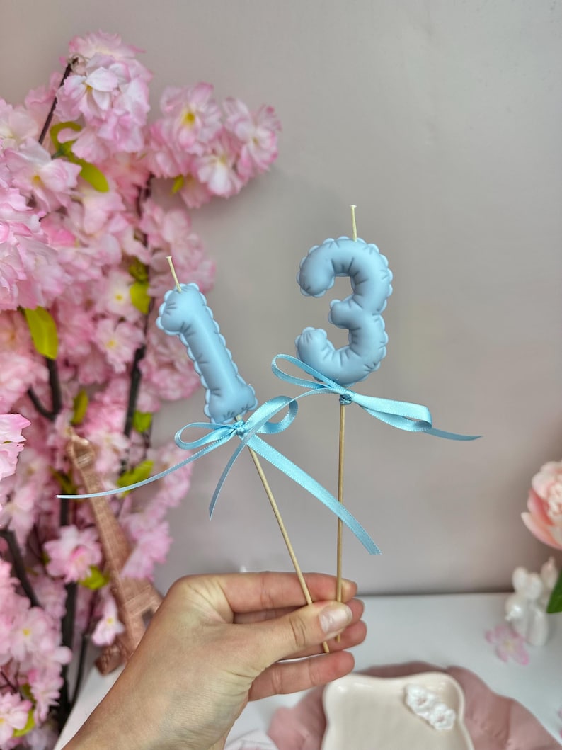 May include: Two light blue number candles, shaped like the digits one and three, are mounted on wooden sticks. Each candle is decorated with a matching light blue satin ribbon tied in a bow around the base of the number.