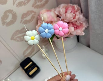 Puede incluir: Tres velas con forma de flor sobre palitos de madera, que incluyen una flor blanca con centro amarillo, una flor azul claro con centro blanco y una flor rosa con centro blanco. Cada vela tiene una mecha que sobresale del centro superior.