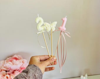 May include: Three decorative cake toppers on wooden sticks: a pale yellow number two candle, a white textured butterfly, and a pale pink number one candle. Each topper is adorned with thin, matching satin ribbons tied below the figures.