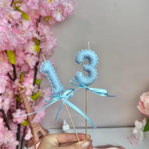 May include: Two light blue number candles, shaped as a one and a three, are mounted on thin wooden sticks. Each candle is decorated with a matching light blue satin ribbon tied in a bow around the base of the number.