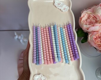 May include: A collection of pastel-colored, beaded candles arranged on a light pink, bear-shaped tray. The candles are in shades of lavender, pink, white, and blue. The tray has white floral embellishments. The candles are a decorative item.